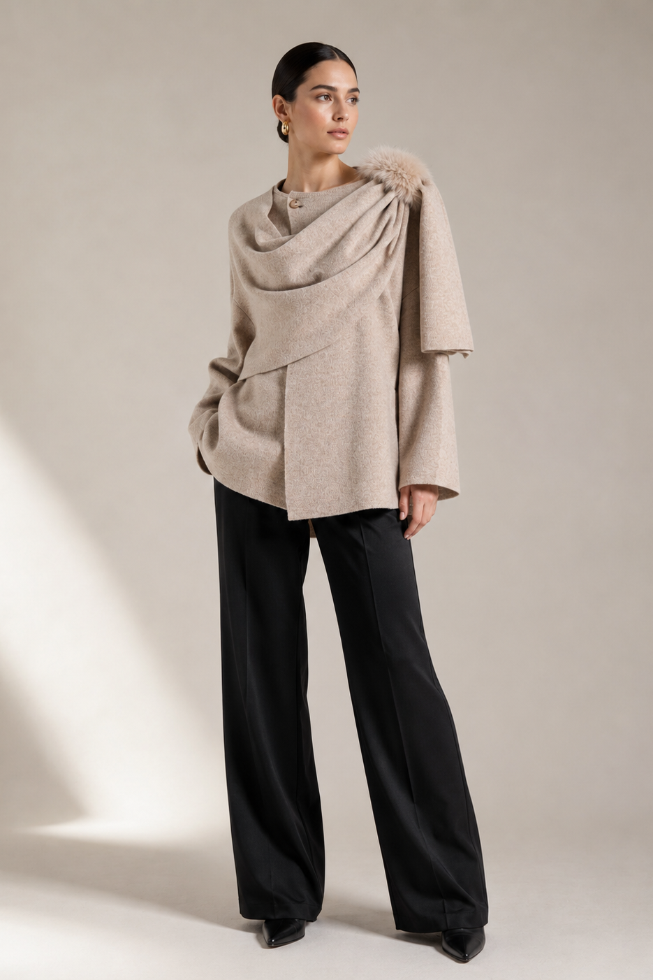 A model stands in a studio wearing a draped, oversized light beige knit top with a fuzzy collar detail, paired with wide-leg black trousers, against a warm beige background with soft diagonal shadows.