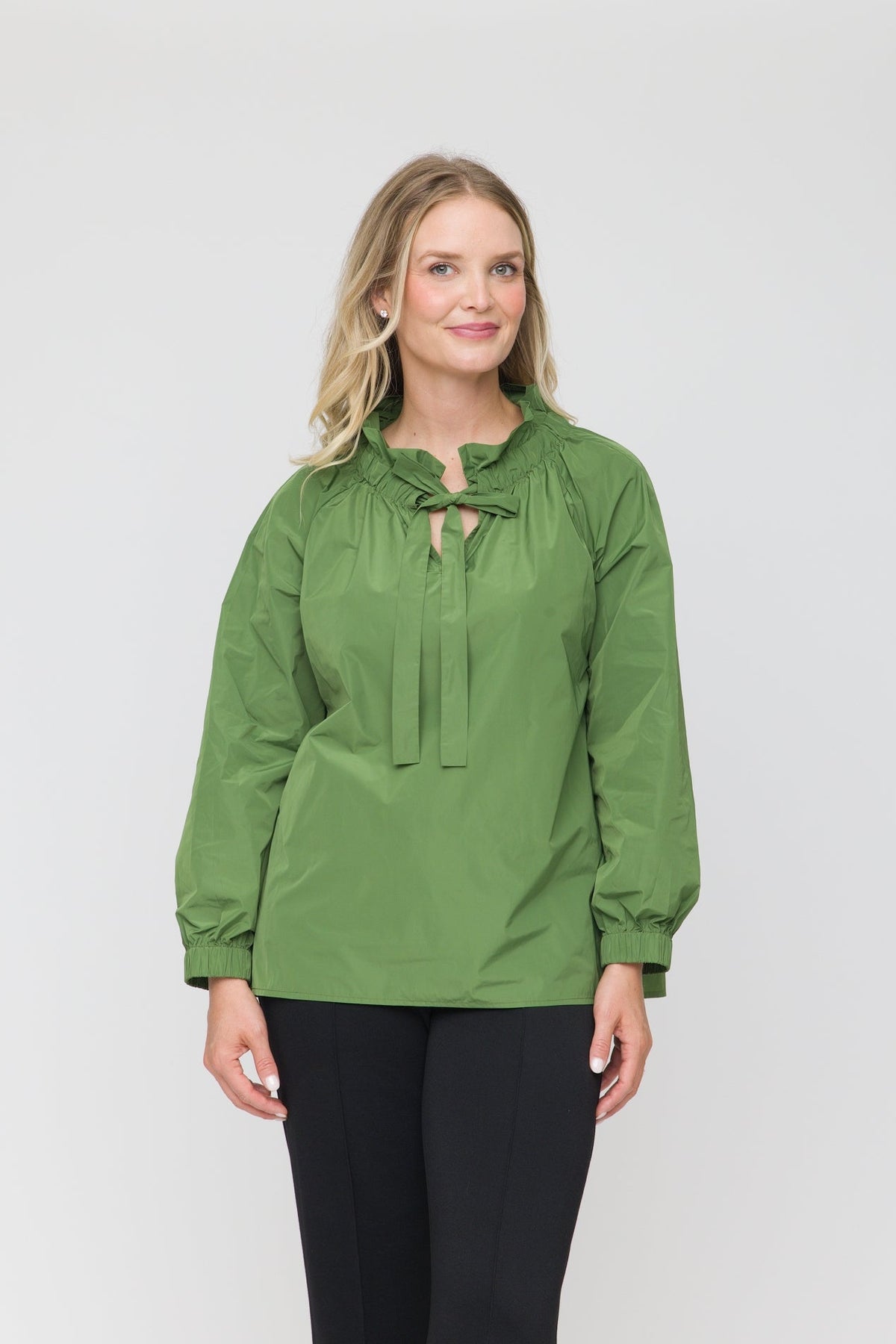A woman modeling a sage green long-sleeve blouse with a tie-neck detail, paired with black pants, against a plain light gray studio background.