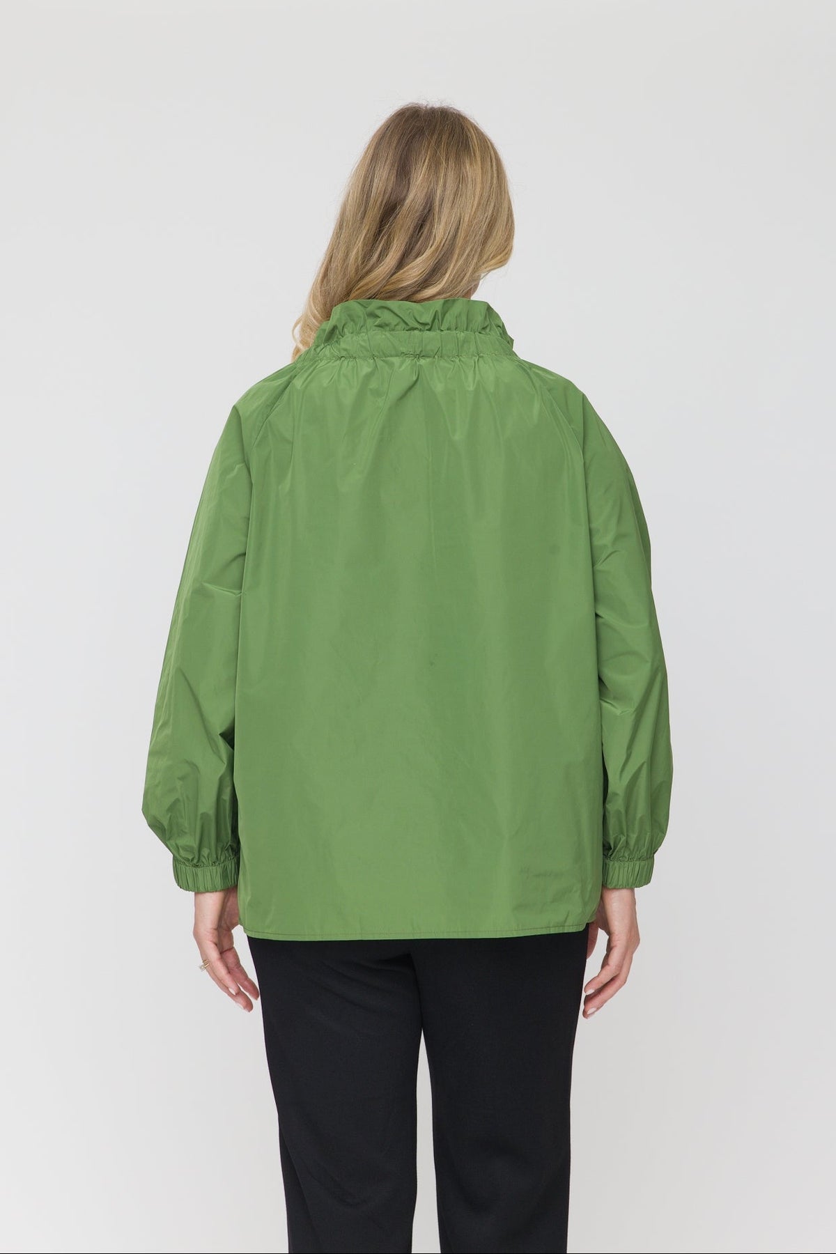 A woman viewed from the back wearing a sage green lightweight jacket and black pants, standing against a plain light gray studio background.