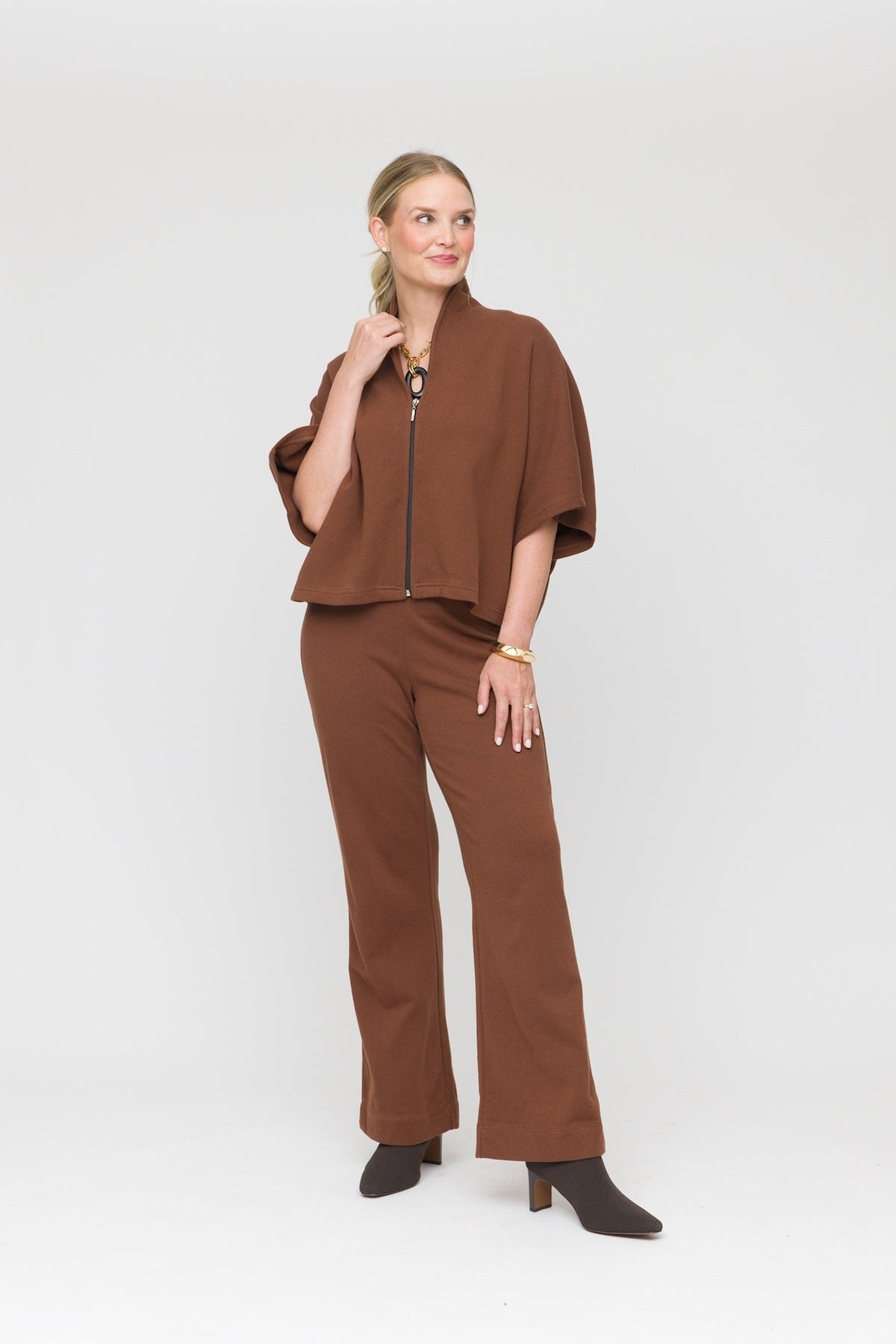 A woman modeling a brown two-piece outfit consisting of a cropped short-sleeve zip-front jacket and matching wide-leg pants, paired with dark brown heeled boots against a light gray studio background.