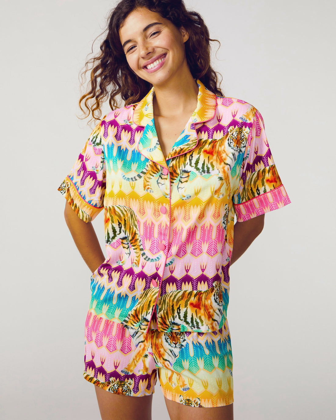 A smiling woman wearing a short-sleeve button-front pajama top and matching shorts in a bright multicolor print with pink, yellow, turquoise, orange, and purple tiger and feather-like motifs, photographed against a light gray studio background.